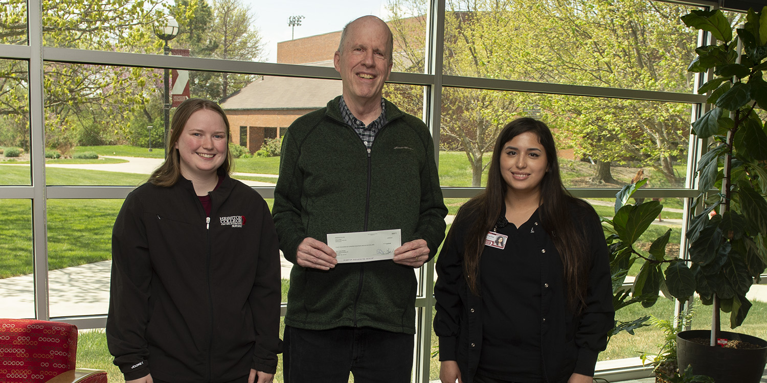 Hesston College nursing students Delaney Lawrence (left) and Michelle Ramos-Carreno (right) present a check to Brian Bisbee, executive director of New Hope Shelter in Newton, in the atrium of Bonnie Sowers Nursing Center.